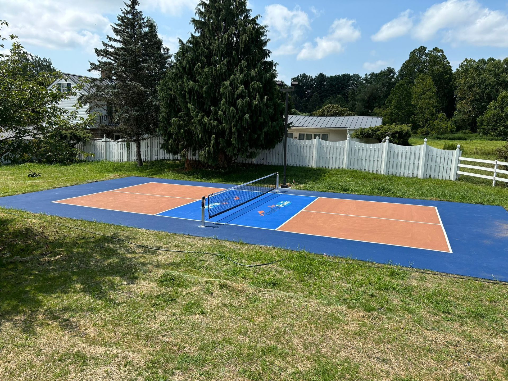 Volleyball court with blue and brown sections, net, on a grassy lawn, white fence in background.