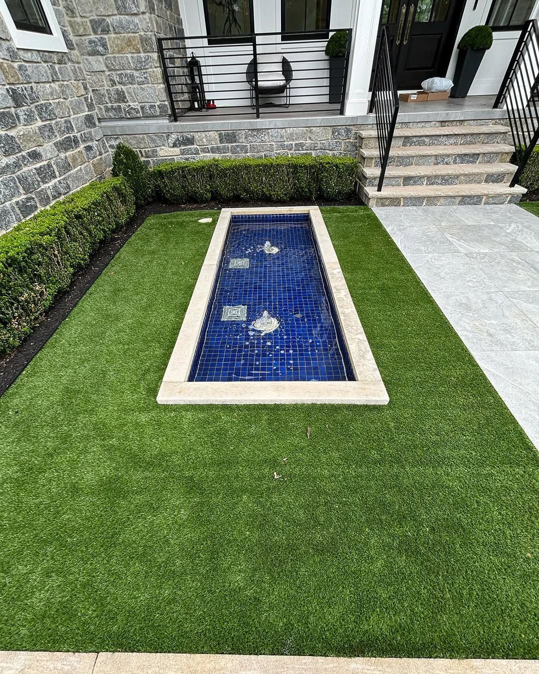 A small pool in the middle of a lush green lawn in front of a house.