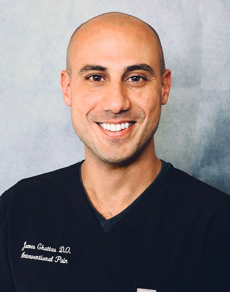Smiling man with shaved head wearing dark scrubs, text on the chest reads