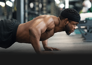 Man doing a push-up in a gym, wearing a black headband and shorts, with blurred equipment in the background