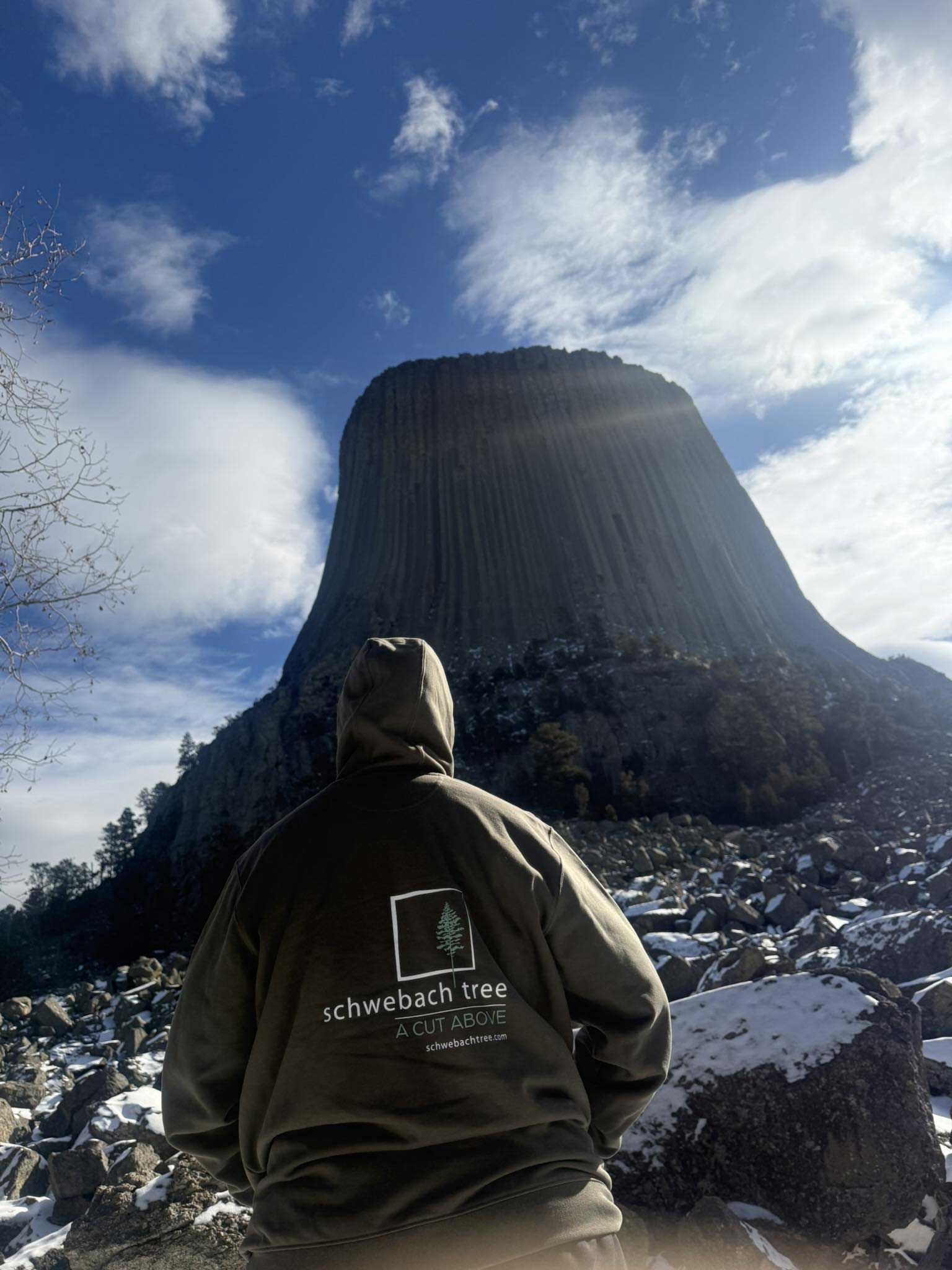 Person in hoodie looks up at Devils Tower under blue sky. Snowy ground.