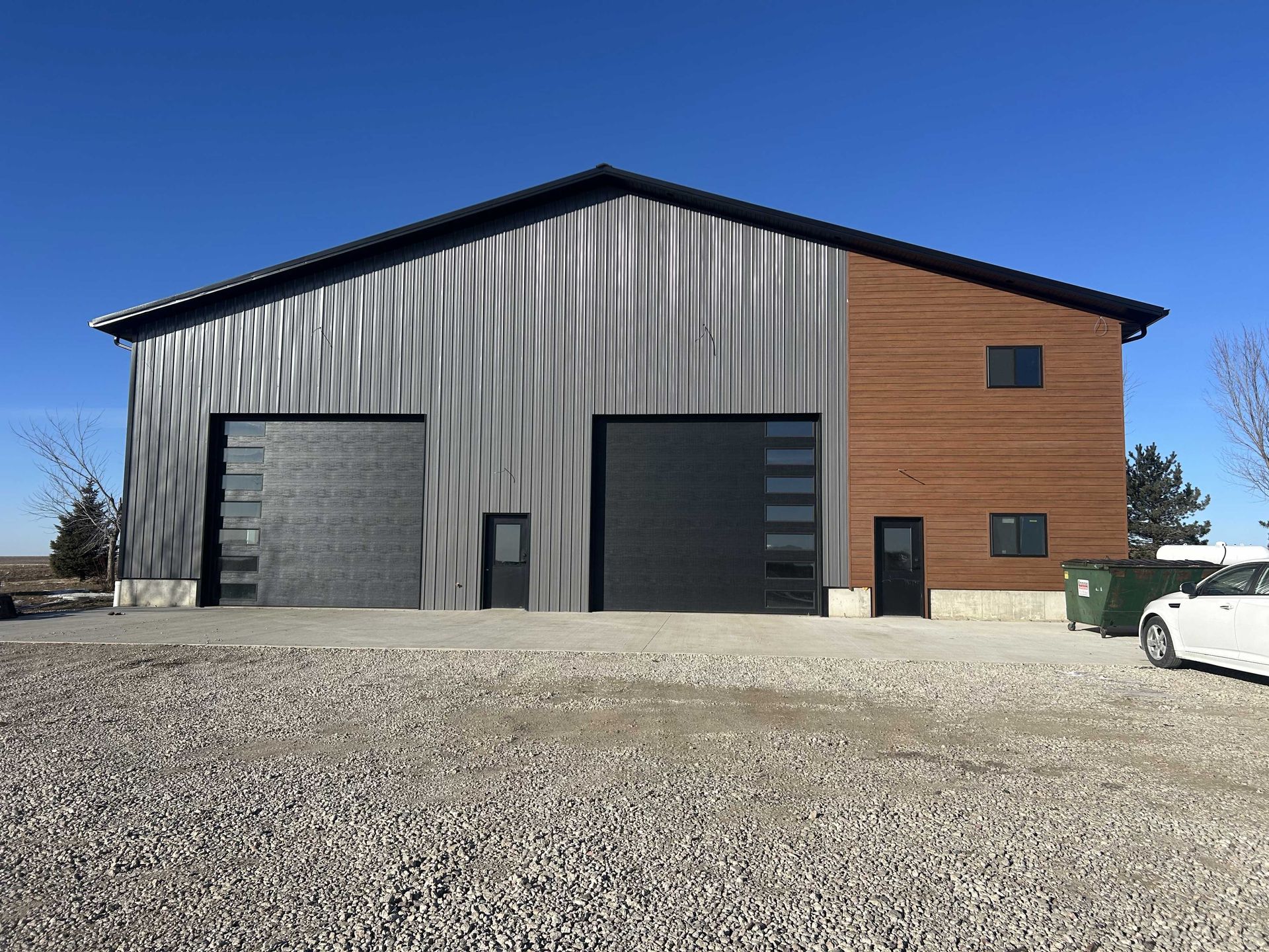 Gray and brown metal building with two garage doors and a gravel driveway under a blue sky.
