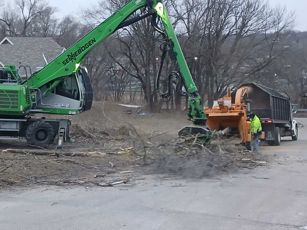 Green machine processing brush, feeding a wood chipper and truck, outdoors.