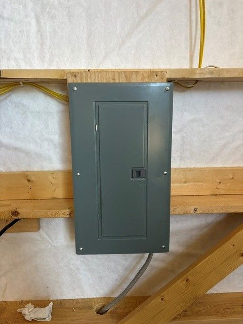 Gray electrical breaker panel mounted on a wooden wall with exposed wiring, in a basement or utility area