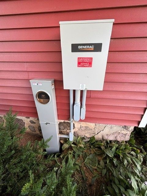 Exterior electrical service boxes mounted on a red siding wall above plants