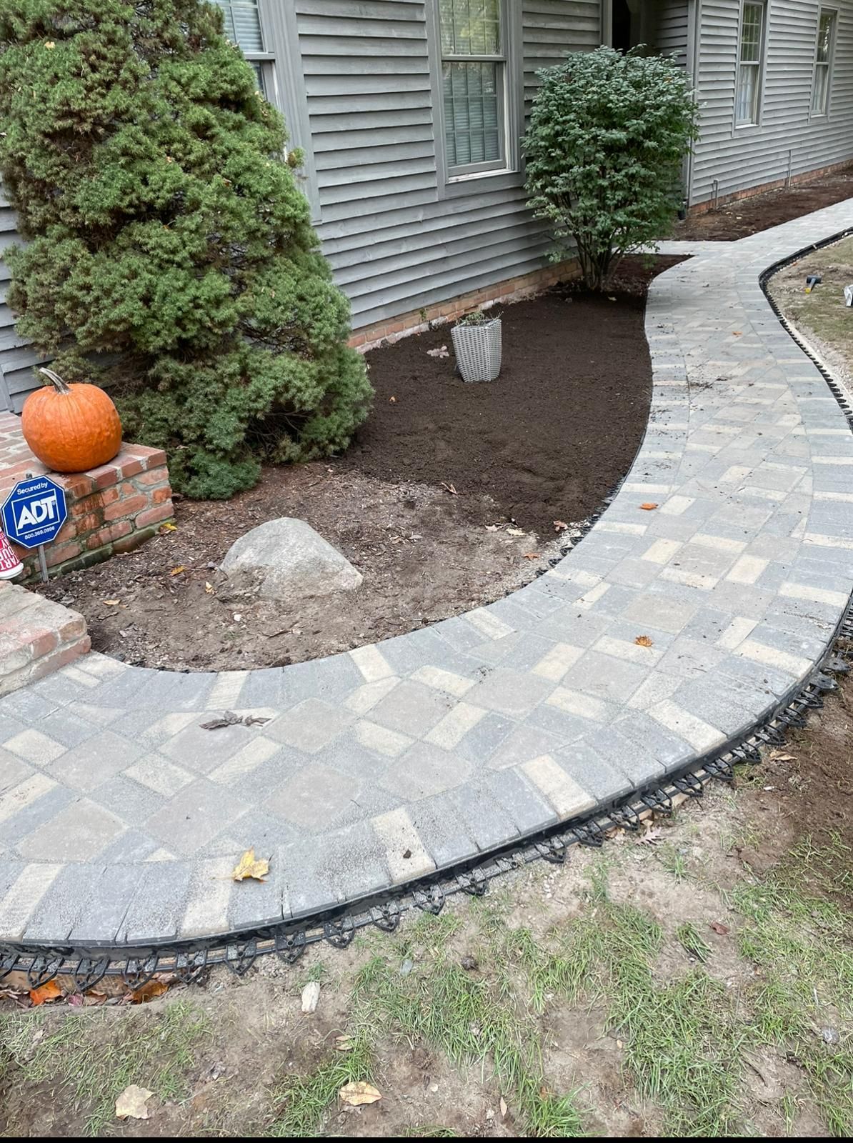 A curved walkway made of grey patterned pavers leads to a house entrance, bordered by landscaping and a mulch garden bed.