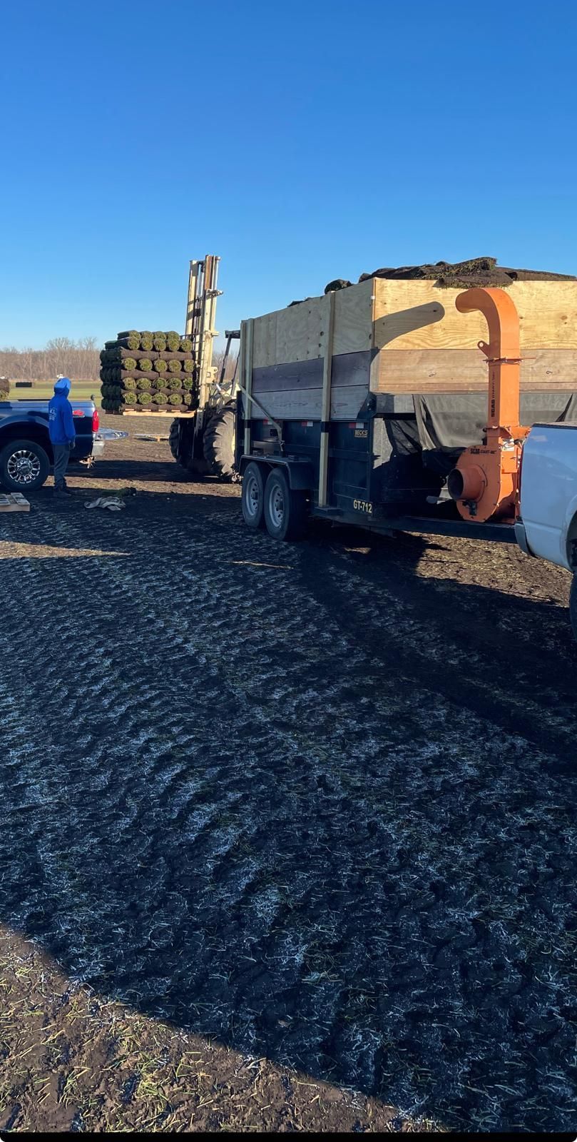 A forklift moves a pallet of sod near a trailer with a leaf blower attachment on a clear day at a sod farm.