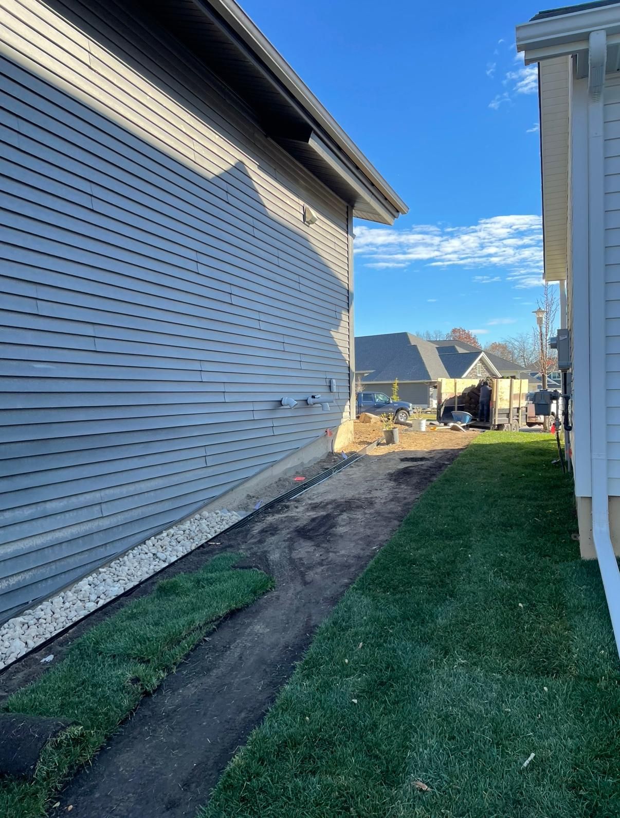 A side yard between two houses showing a strip of bare dirt and gravel leading to a sunny backyard.