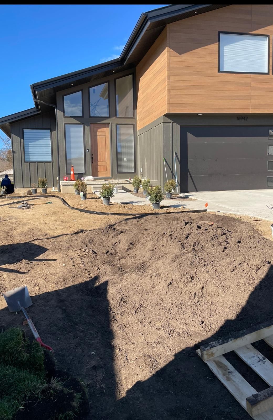 A modern home with wood siding and a driveway, featuring a freshly excavated dirt patch in the front yard.