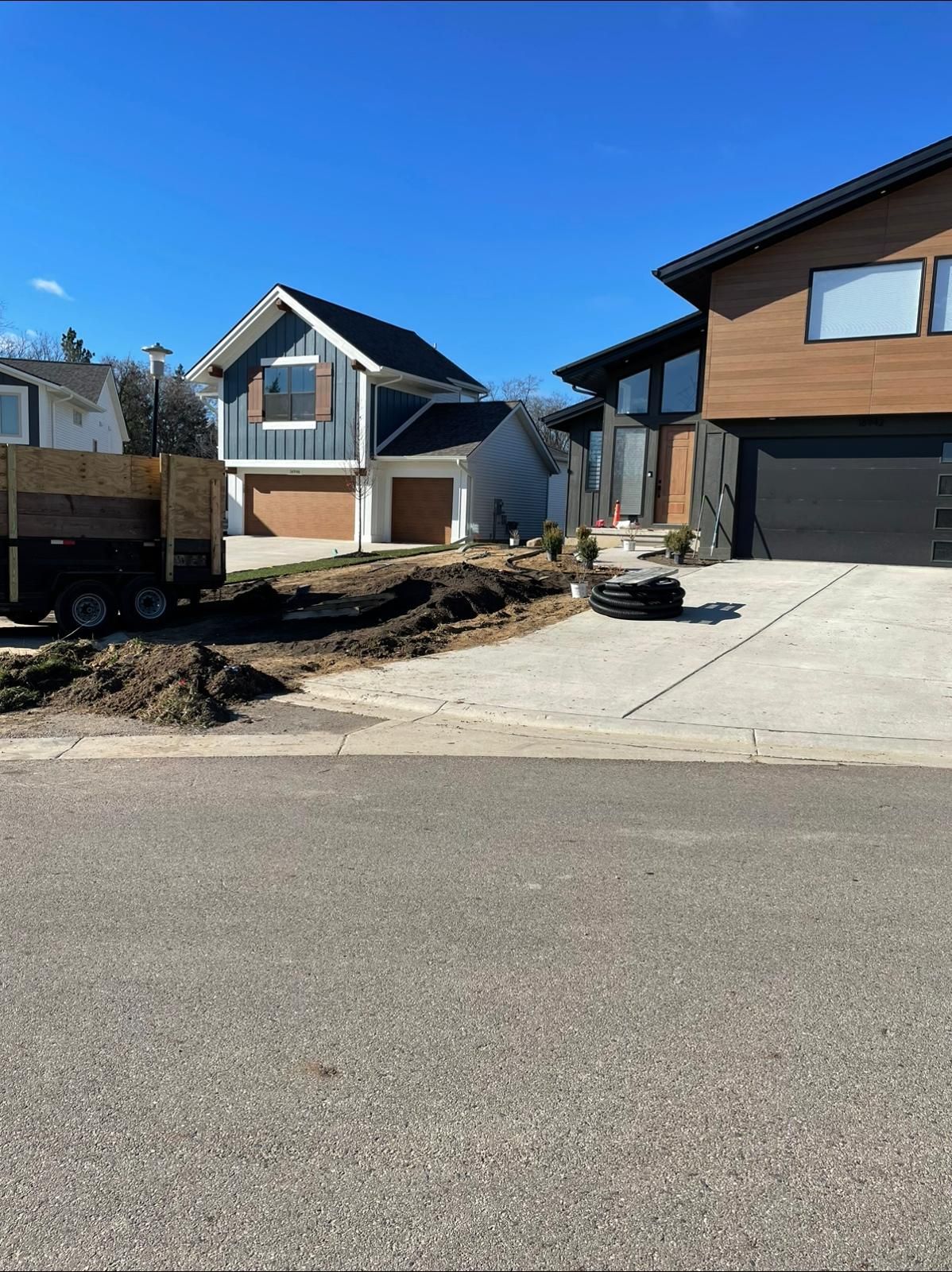 Two modern suburban houses under a clear blue sky, with a dump trailer parked in the foreground by an unlandscaped yard.