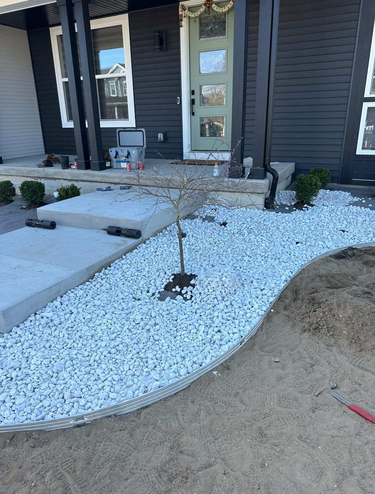 A newly landscaped yard with white decorative stones, a small sapling, and metal edging in front of a dark-sided house.