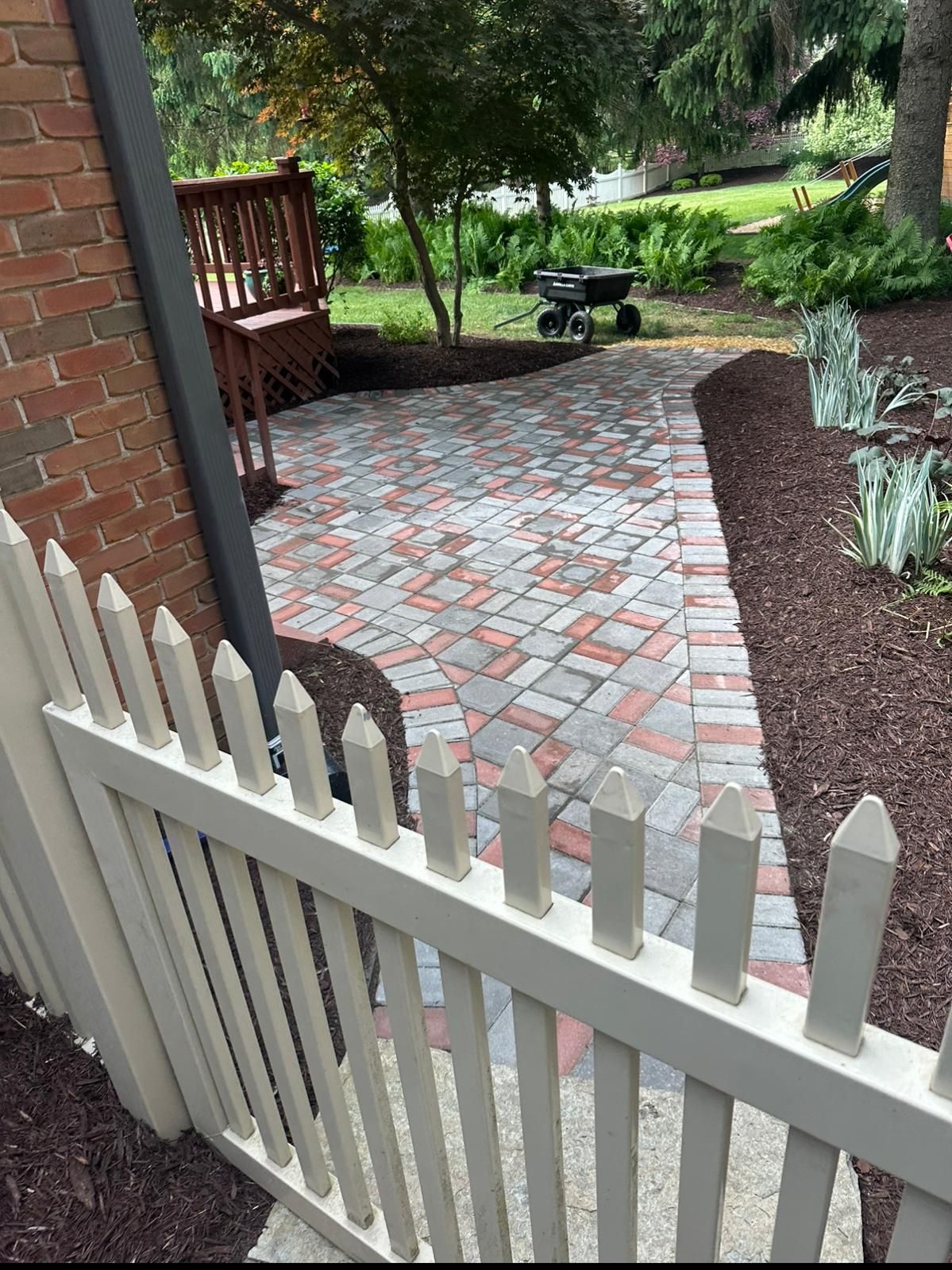 A paved patio with a mix of red and grey bricks leads to a wooden deck, bordered by mulch, plants, and a white picket fence.