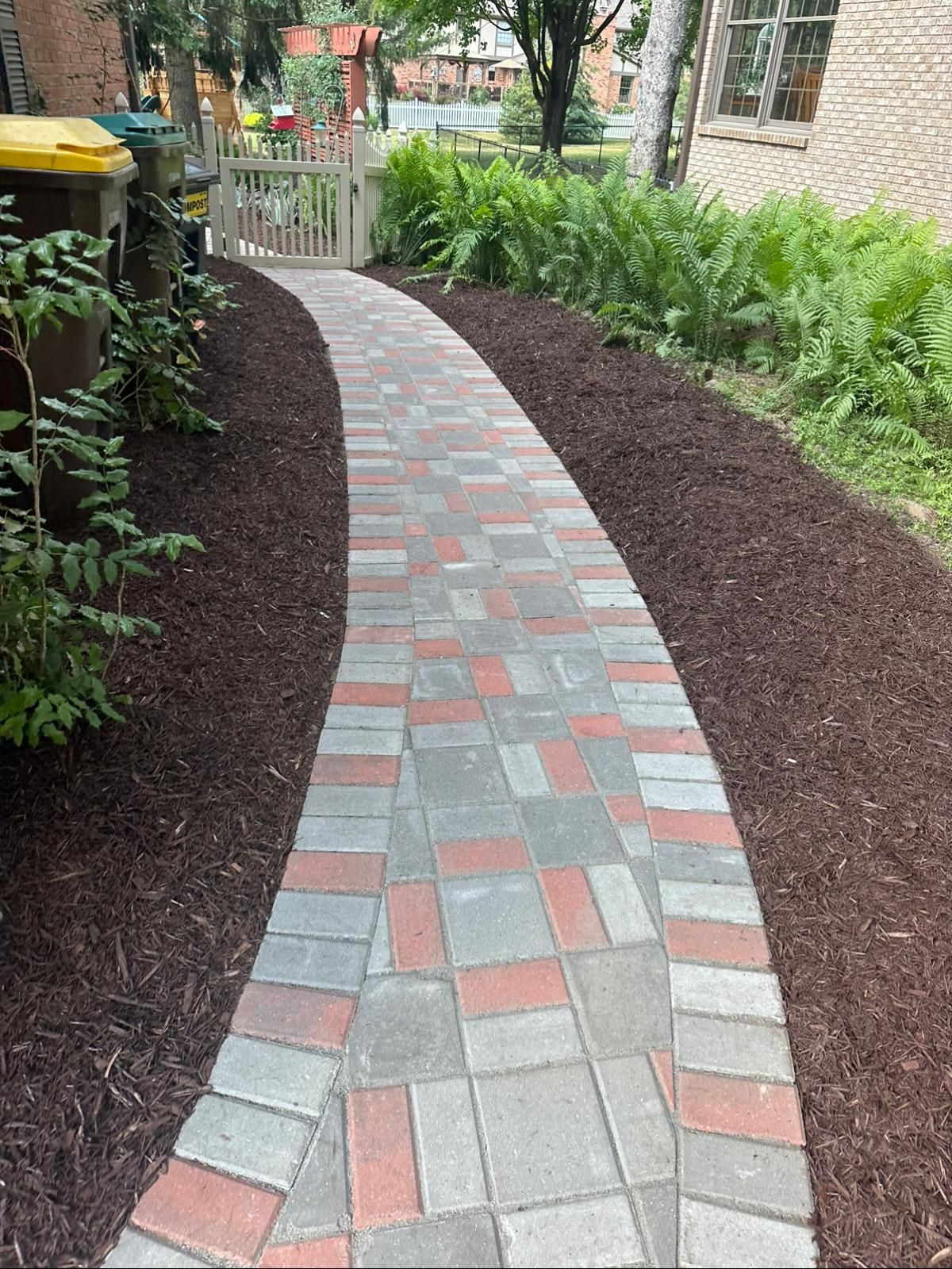 A curved walkway made of grey and red pavers, lined with dark mulch and green ferns leading toward a white gate.