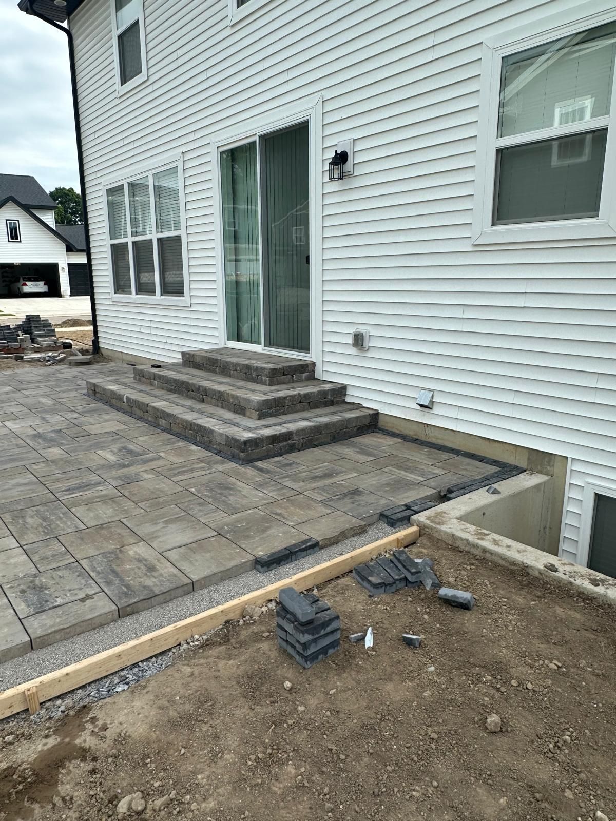 A new grey stone patio with built-in stairs leading to a white house, with an unfinished yard area in the foreground.