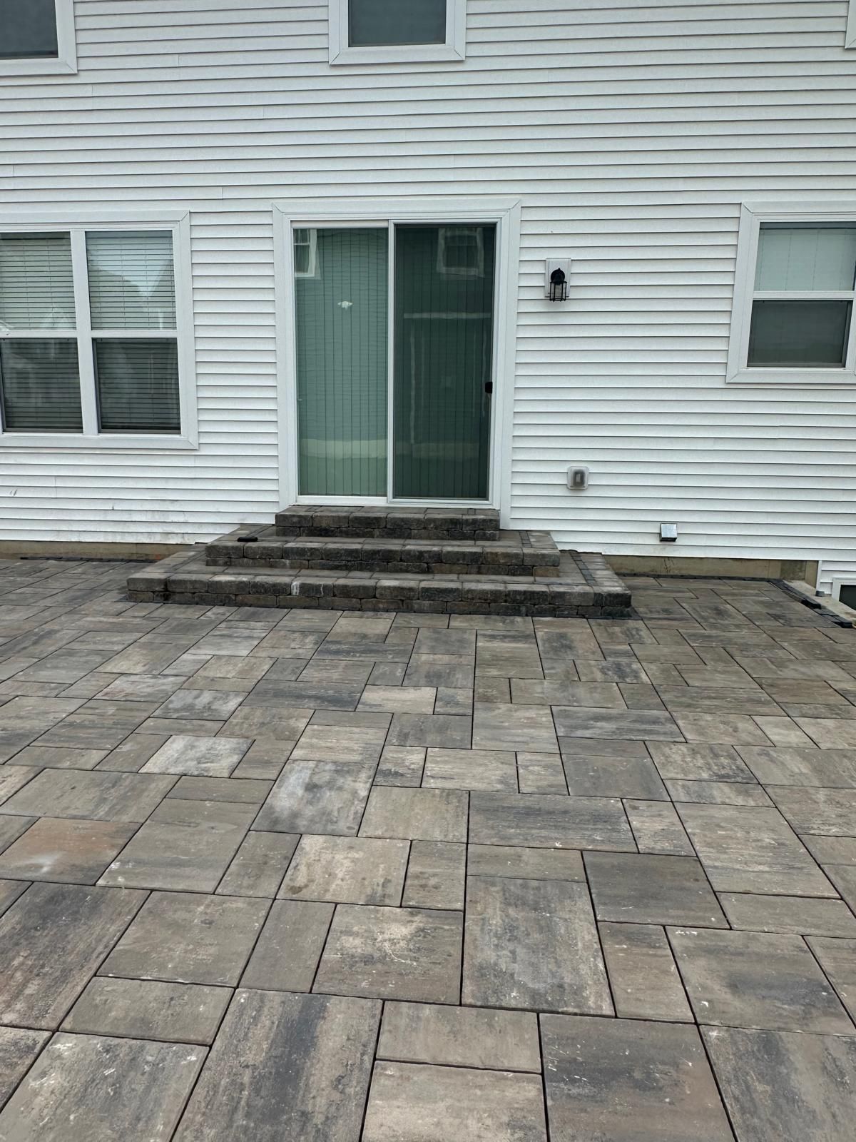 A light-colored house with white siding and a sliding glass door leading to stone steps and a paved patio.