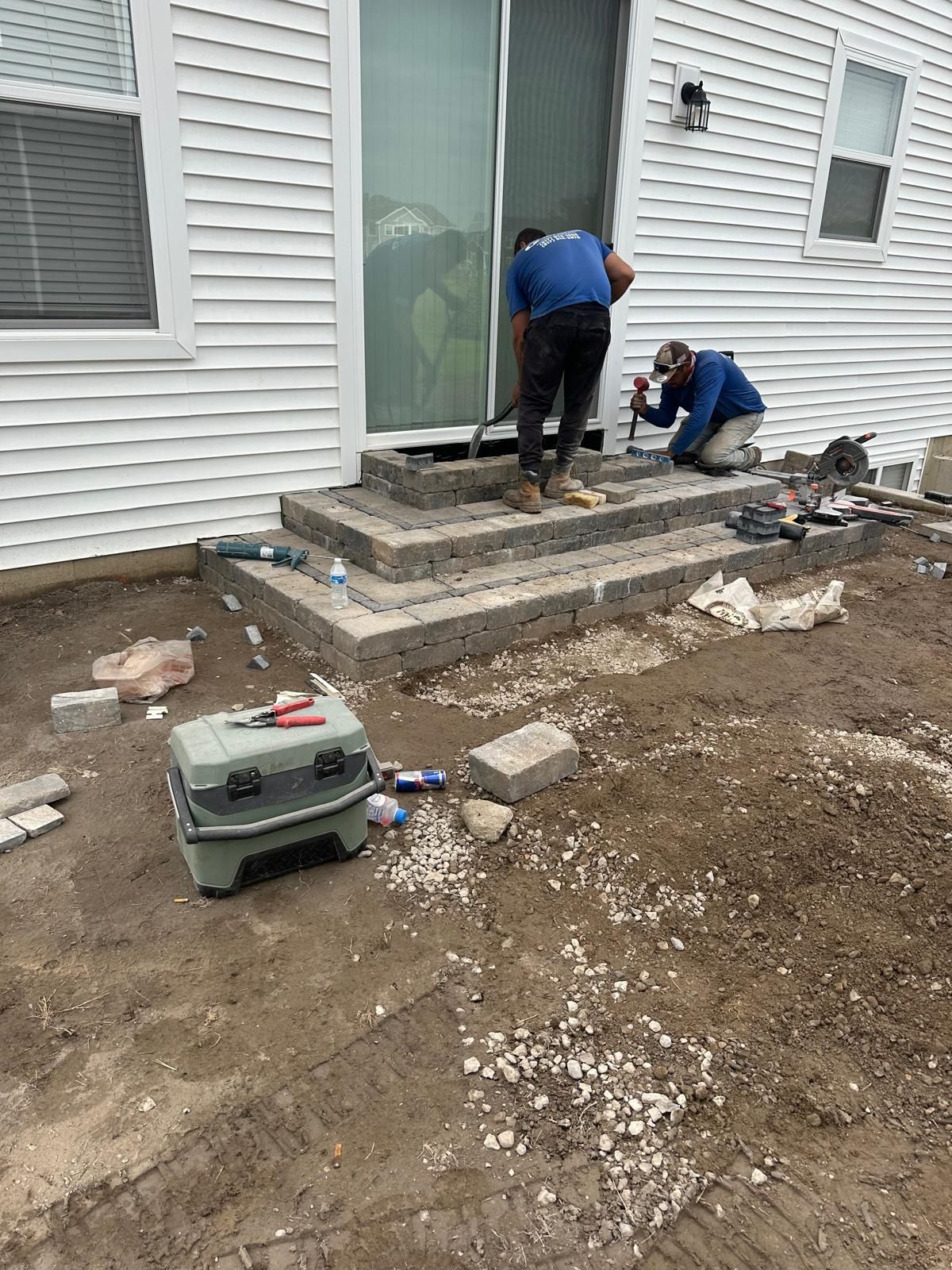 Two workers building stone steps in front of a white house with a sliding glass door.