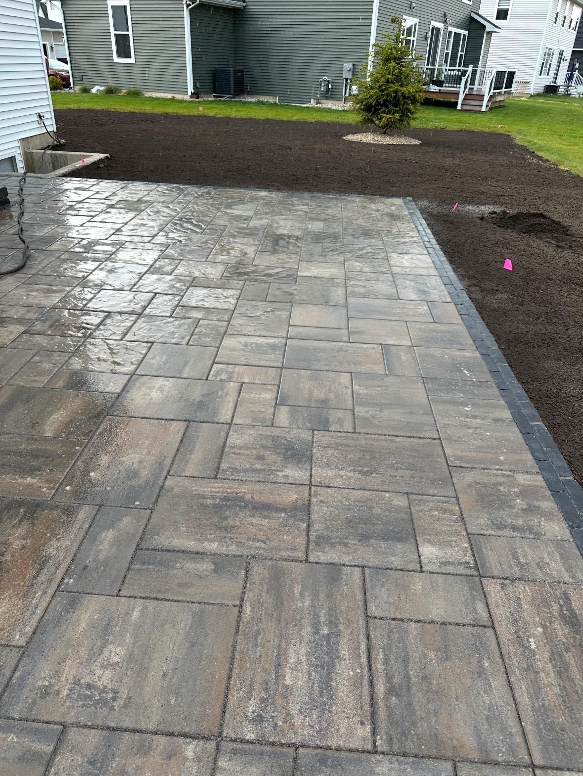 A new rectangular paver patio with a dark border sits next to a residential home and an area of fresh topsoil.