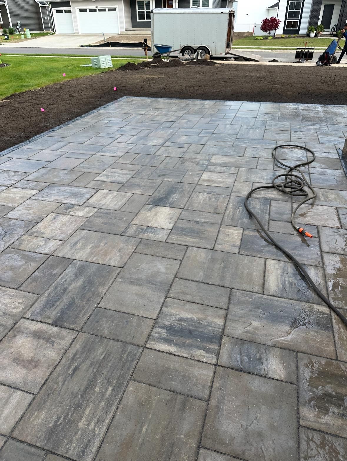 A newly installed patio made of grey rectangular pavers, with a hose coiled on the surface and a yard in the background.
