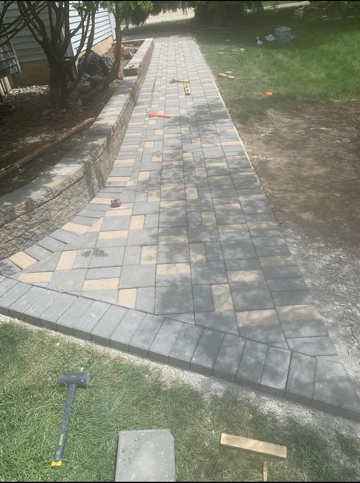 A freshly installed gray and tan paver walkway with a soldier course border next to a stone retaining wall on grass.