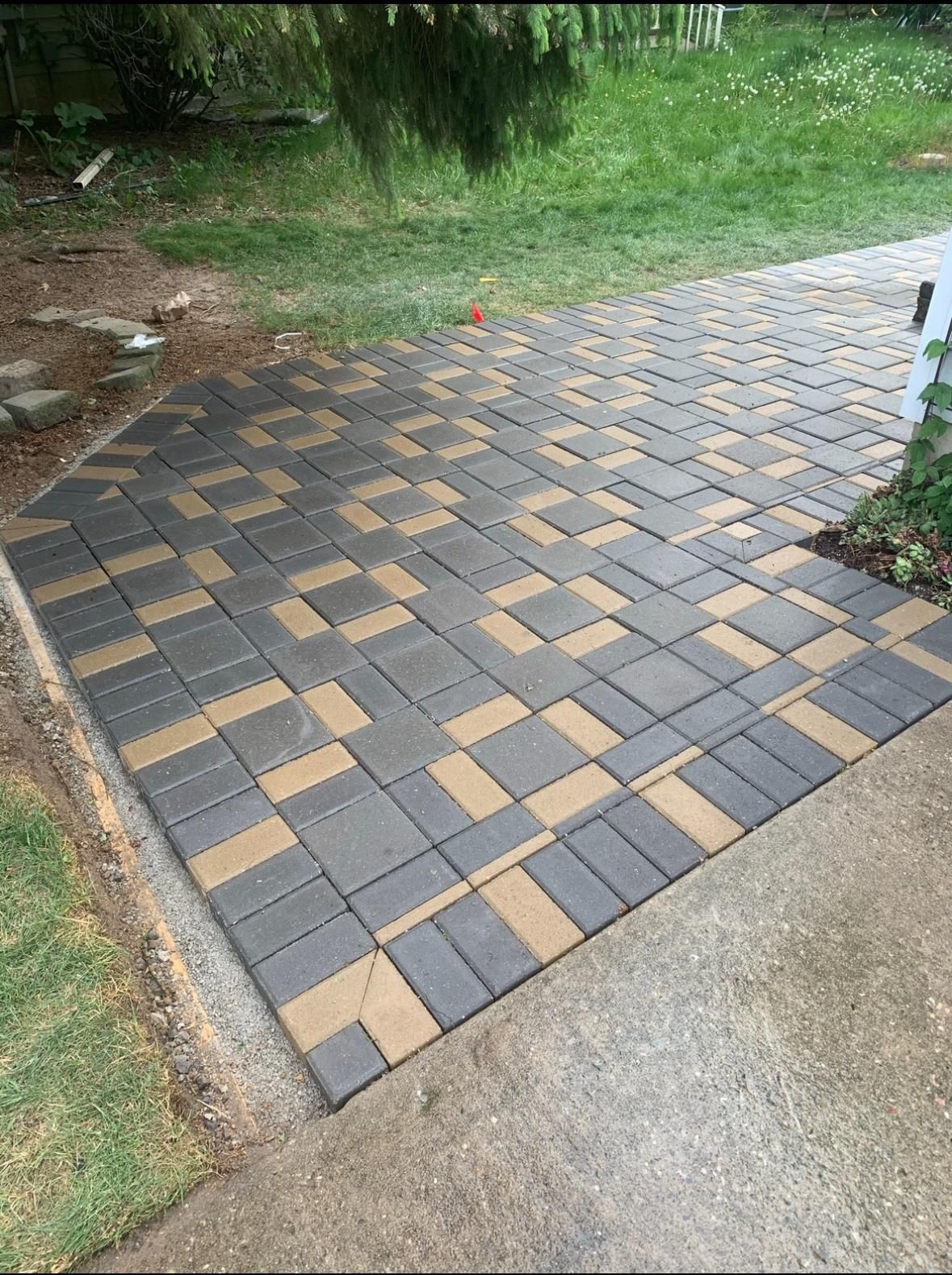 A modern patio paved with interlocking rectangular stones in a mix of dark gray and tan, set against a grassy yard edge.