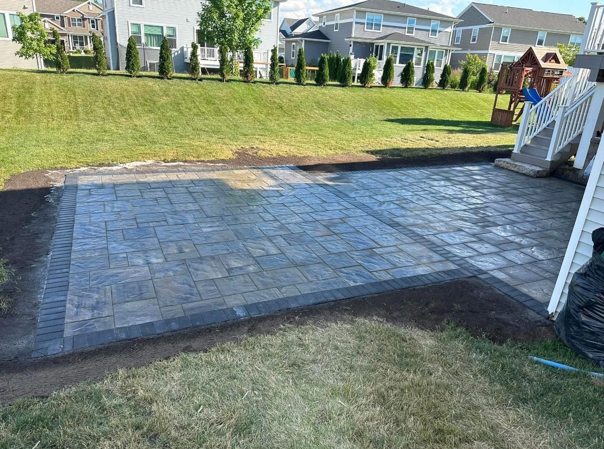 A newly installed rectangular paver patio with a dark border, positioned in a grassy backyard next to a house deck.