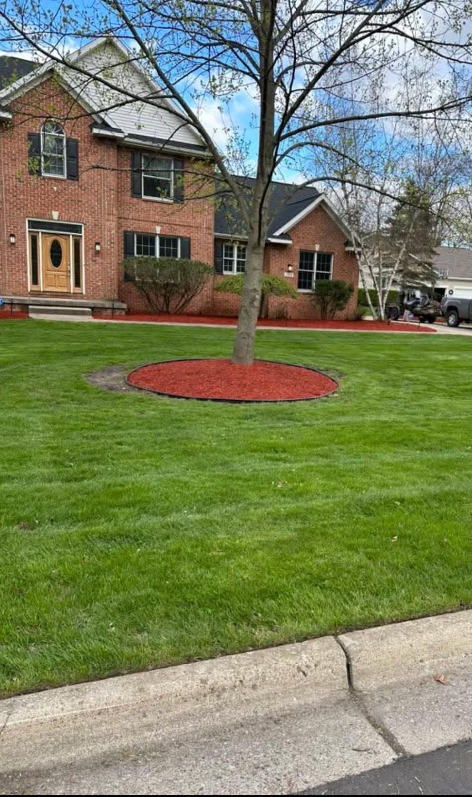 A two-story brick house with a green lawn, a tree with a red mulch ring, and red landscape edging.