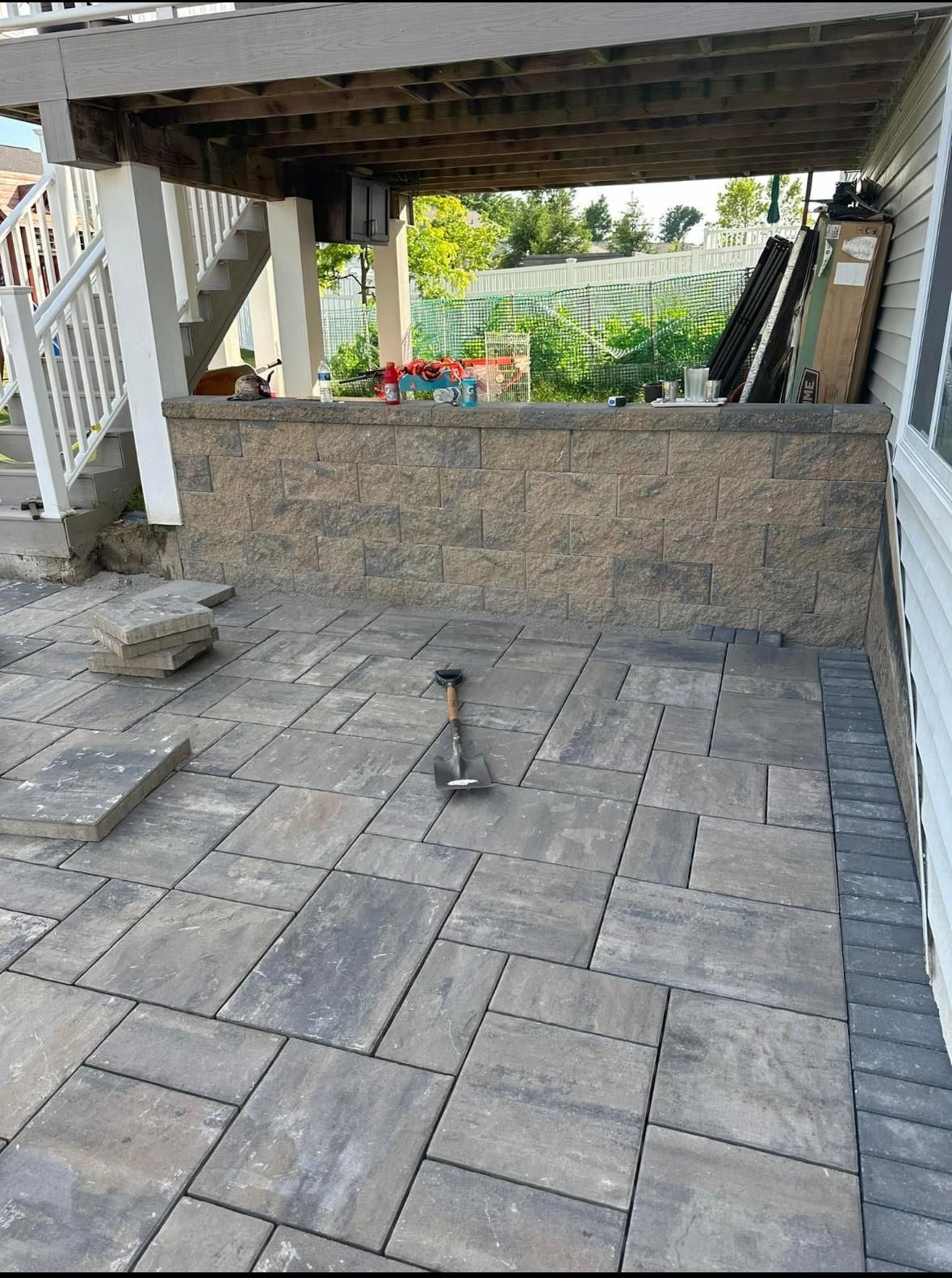 A stone patio under a deck with a retaining wall, a shovel lying in the center, and a stairwell on the left.