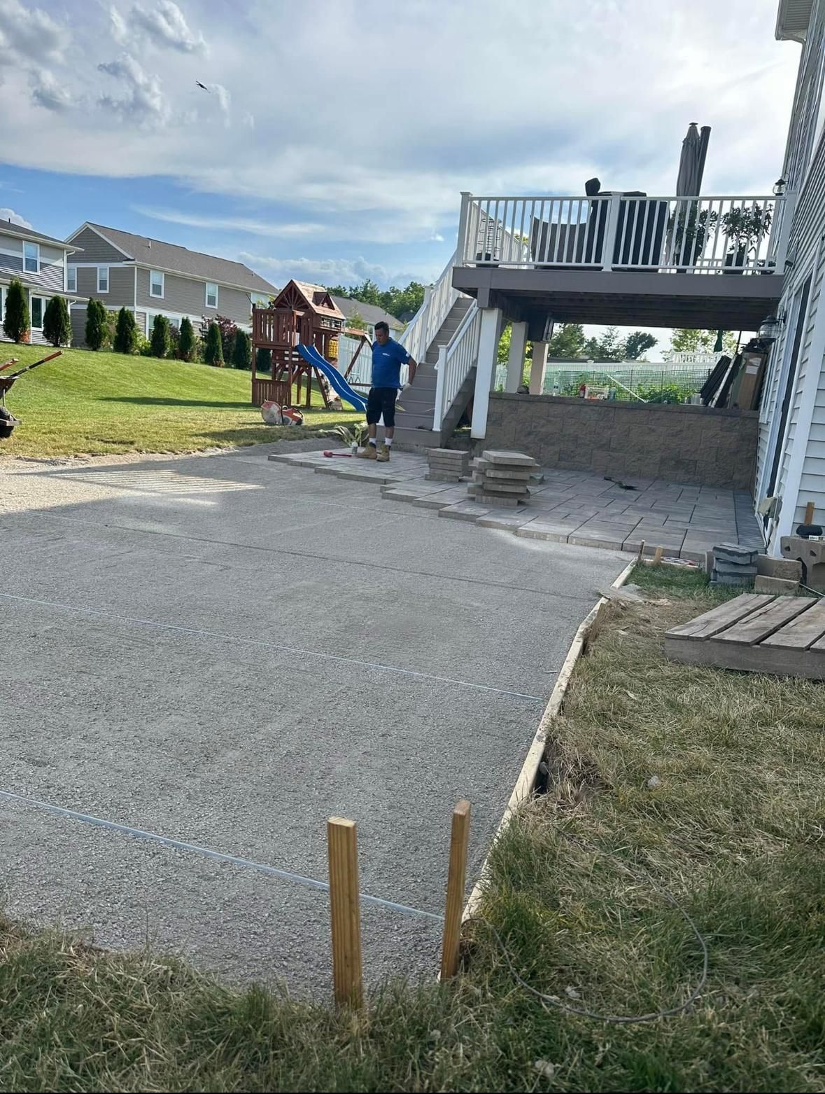 A person works on a paver patio construction project under an elevated deck in a residential backyard.