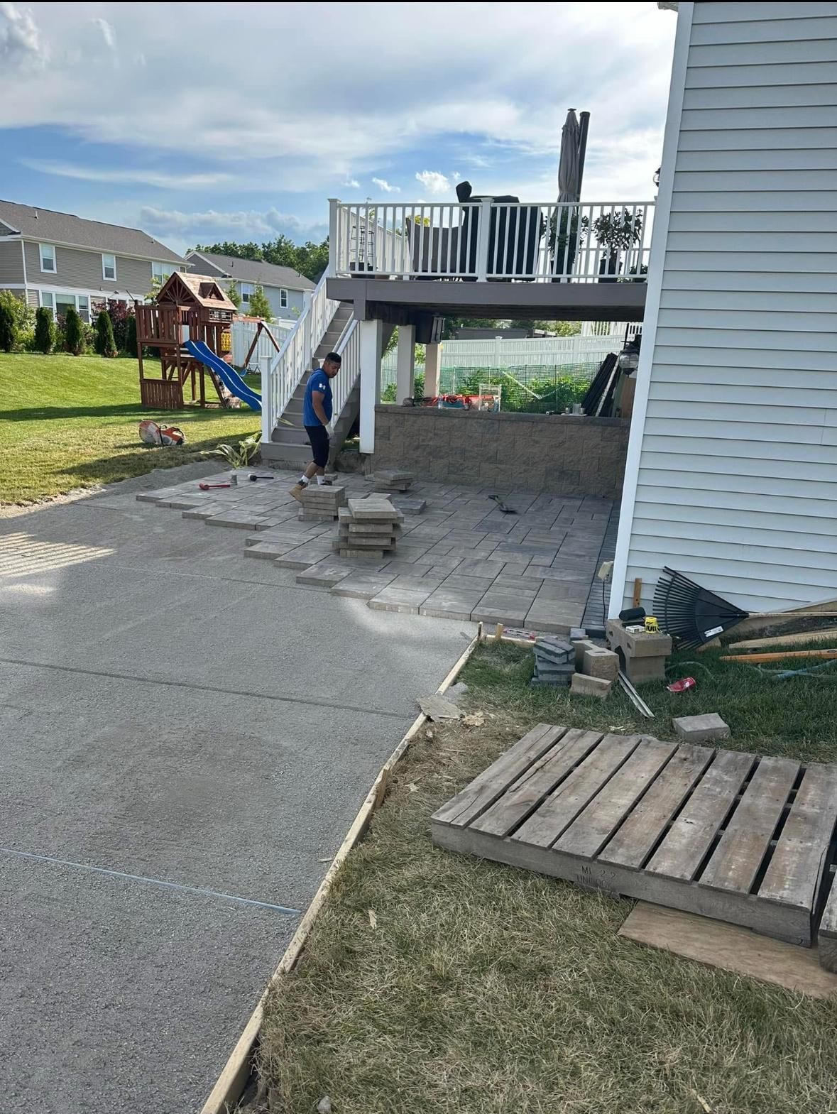 A person laying patio pavers under an elevated wooden deck in a backyard with a house and play set nearby.
