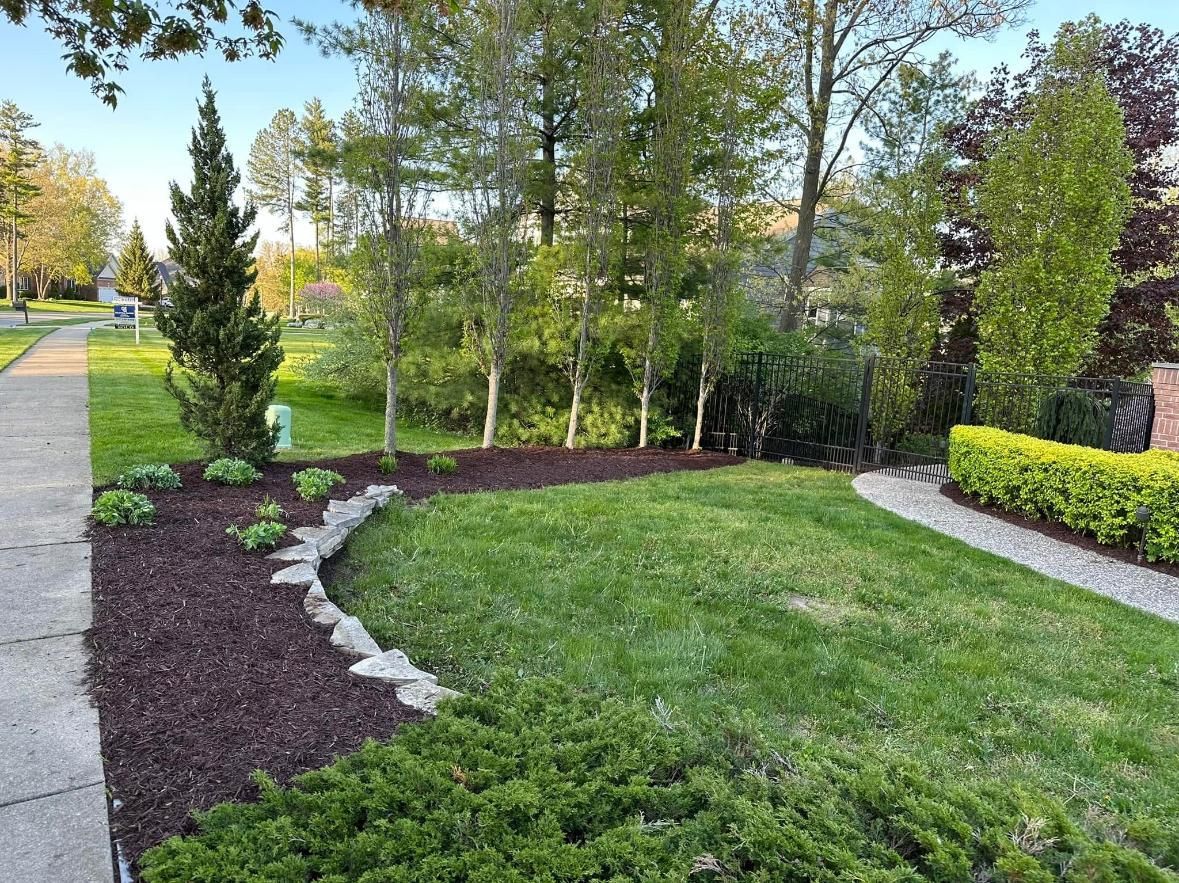 A suburban front yard features a manicured lawn, a curved rock-bordered garden bed with mulch, shrubs, and tall trees.