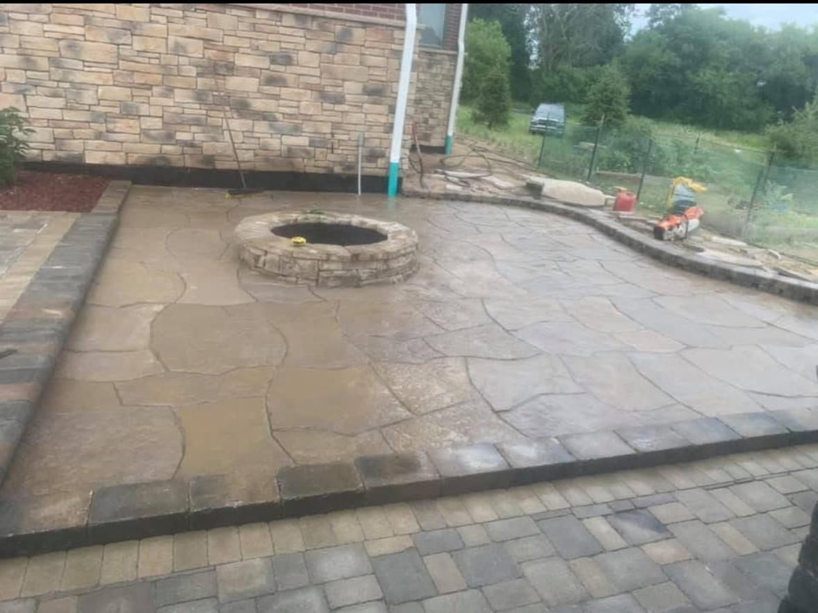 A backyard patio featuring irregular stone pavers, a central circular stone fire pit, and a stone house wall.