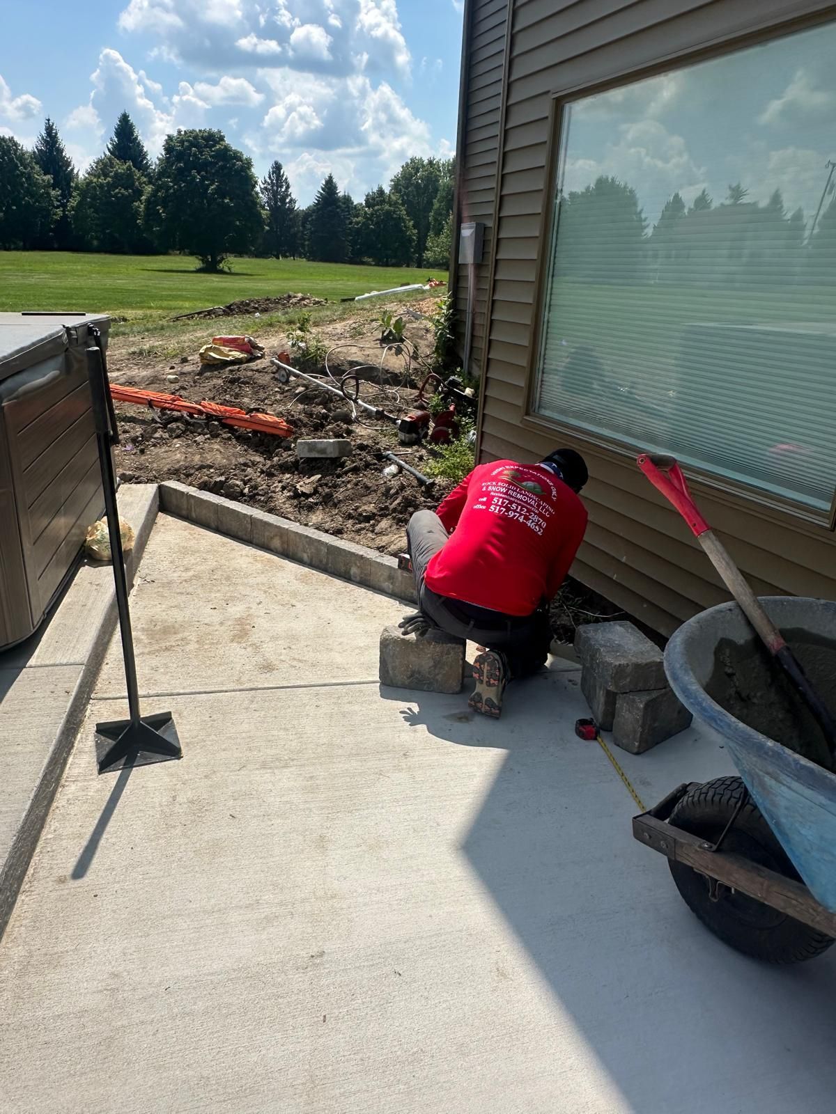 A worker in a red shirt kneels on a concrete patio next to a house, installing concrete blocks near a dirt yard.