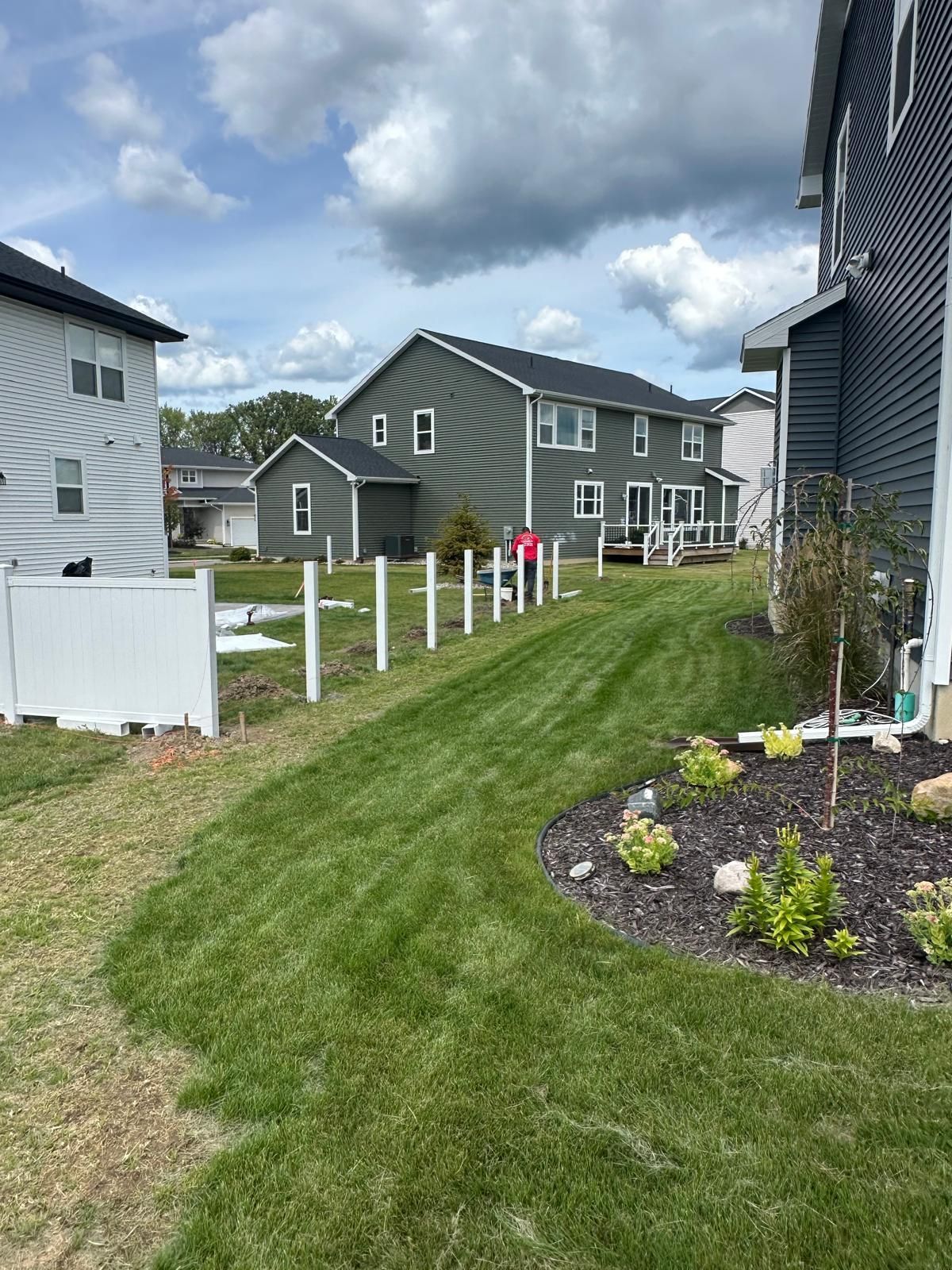 A residential backyard scene featuring newly installed white fence posts between a grey house and a white house.