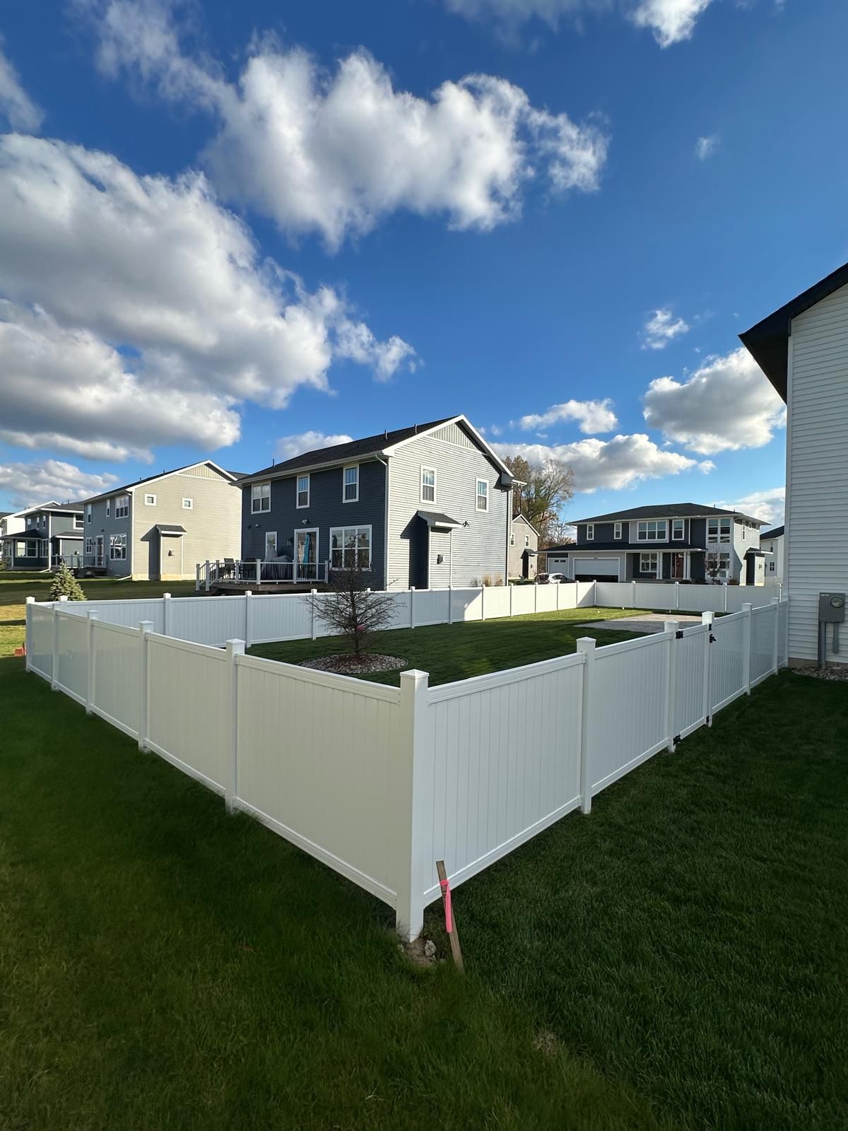 A white vinyl fence encloses a green backyard on a sunny day with houses in the background.