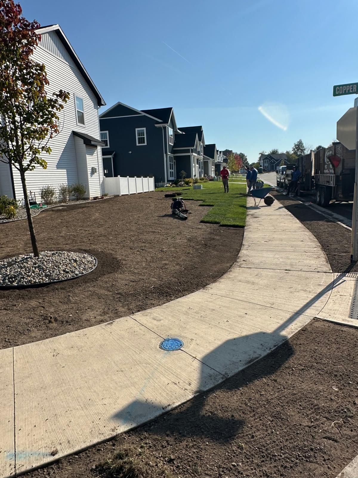 Workers laying sod on a patch of dirt lawn beside a suburban sidewalk and street on a sunny day.