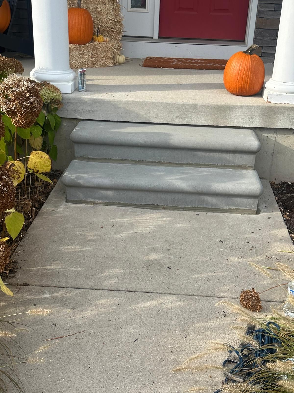 Two concrete steps leading to a front porch decorated with pumpkins, hay, and dried flowers.