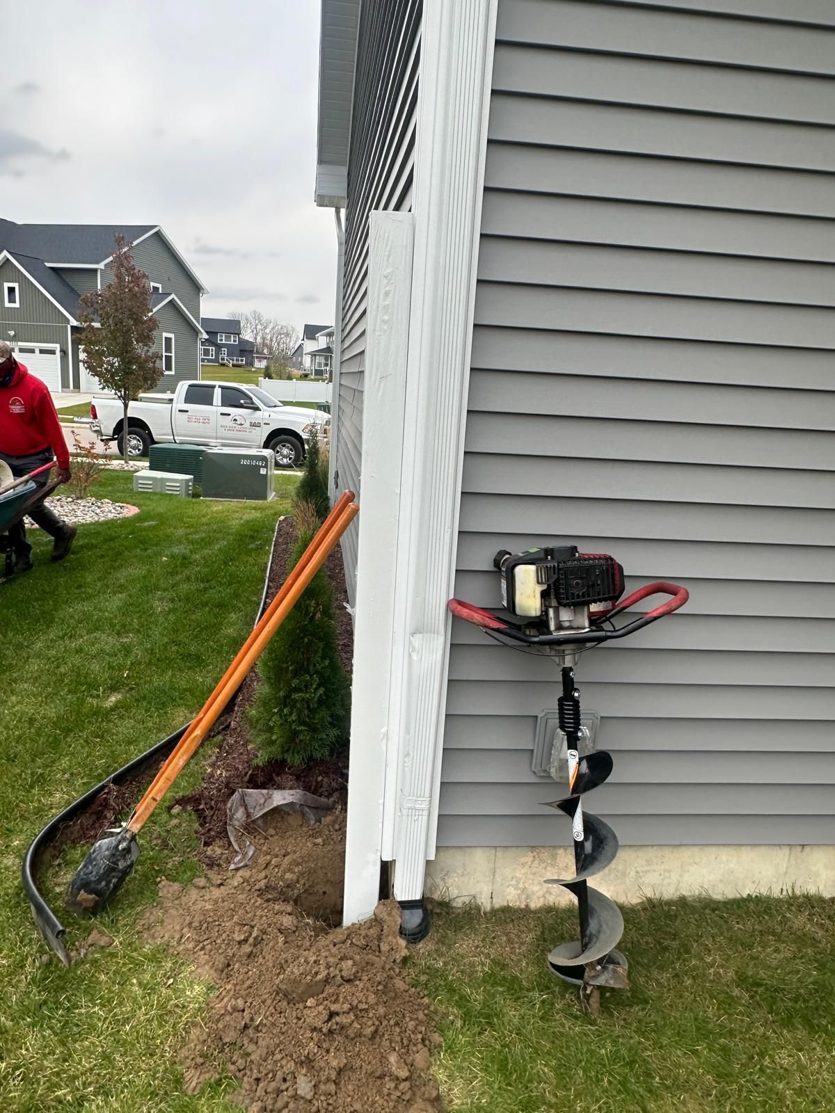 An auger stands next to a gray house siding, with a shovel and fresh dirt nearby in a suburban yard.
