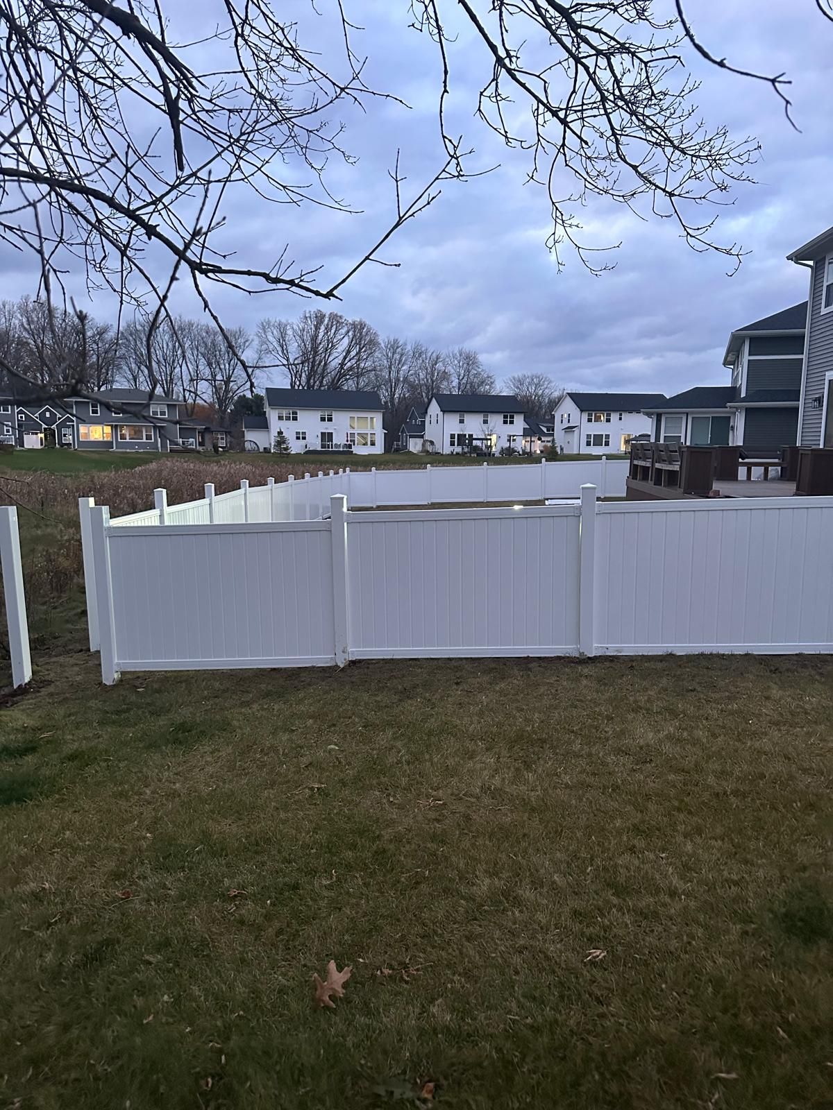 A white vinyl fence stands in a grassy backyard, with residential homes visible in the background under a cloudy sky.