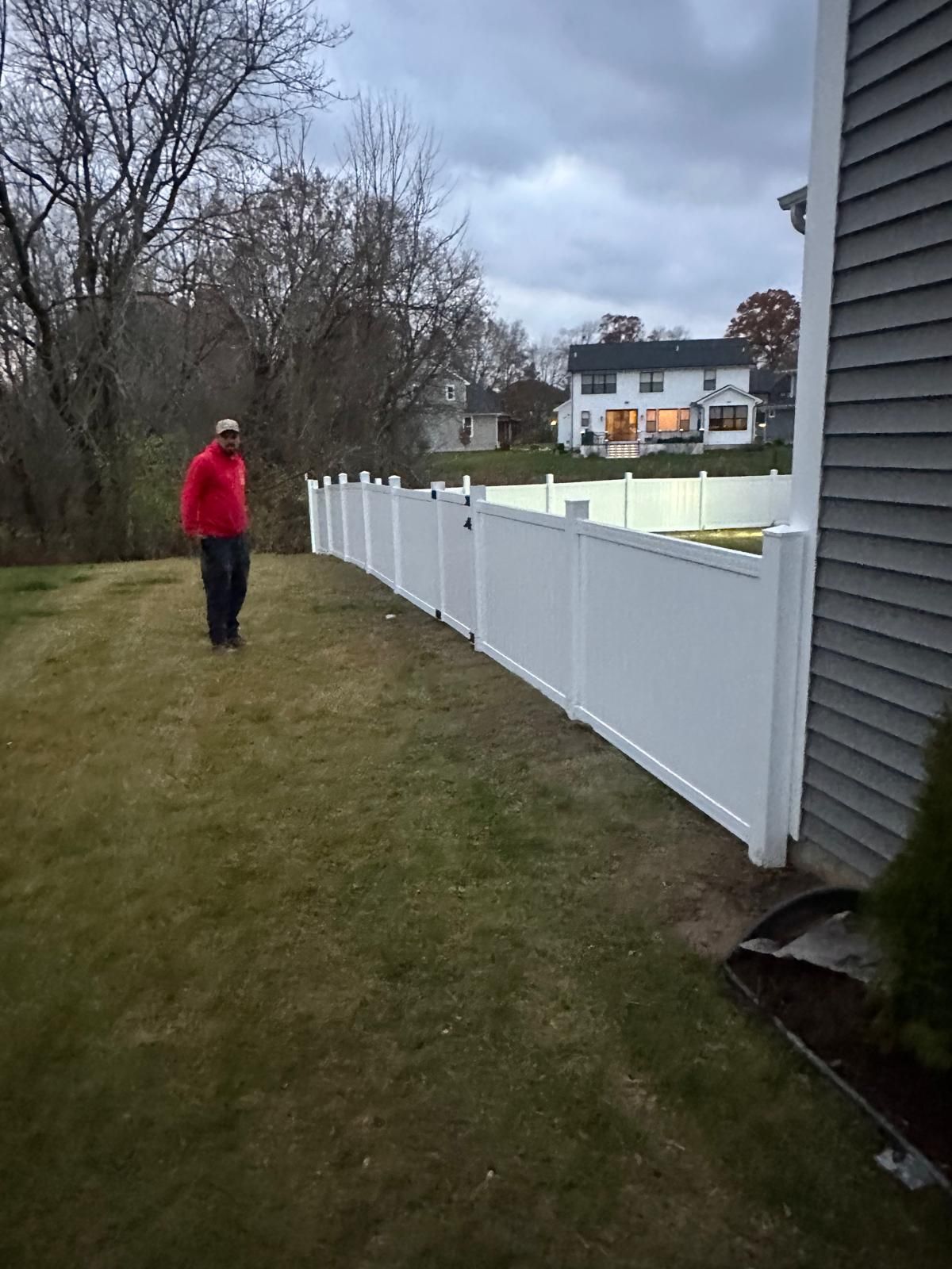 A person in a red jacket stands on a grassy lawn next to a white vinyl fence that extends from the side of a house.
