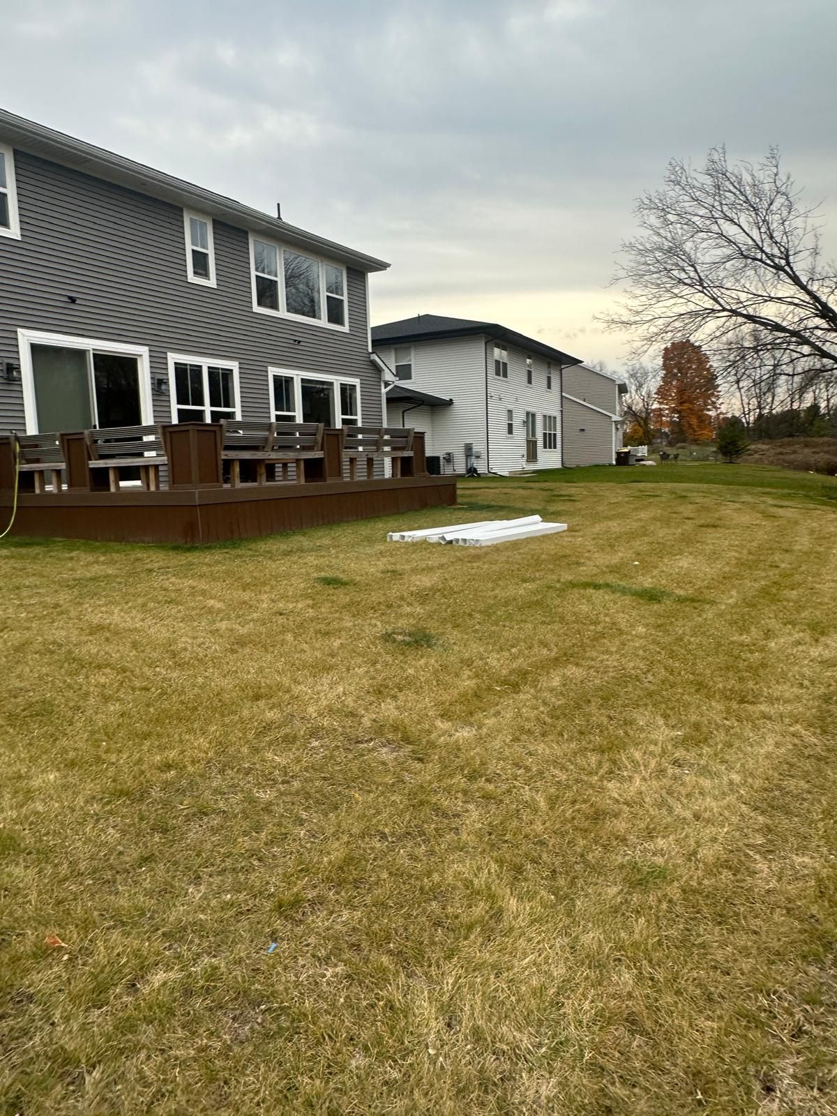A grey two-story house with a wooden deck stands next to a white house on a large, dry grass lawn under a cloudy sky.