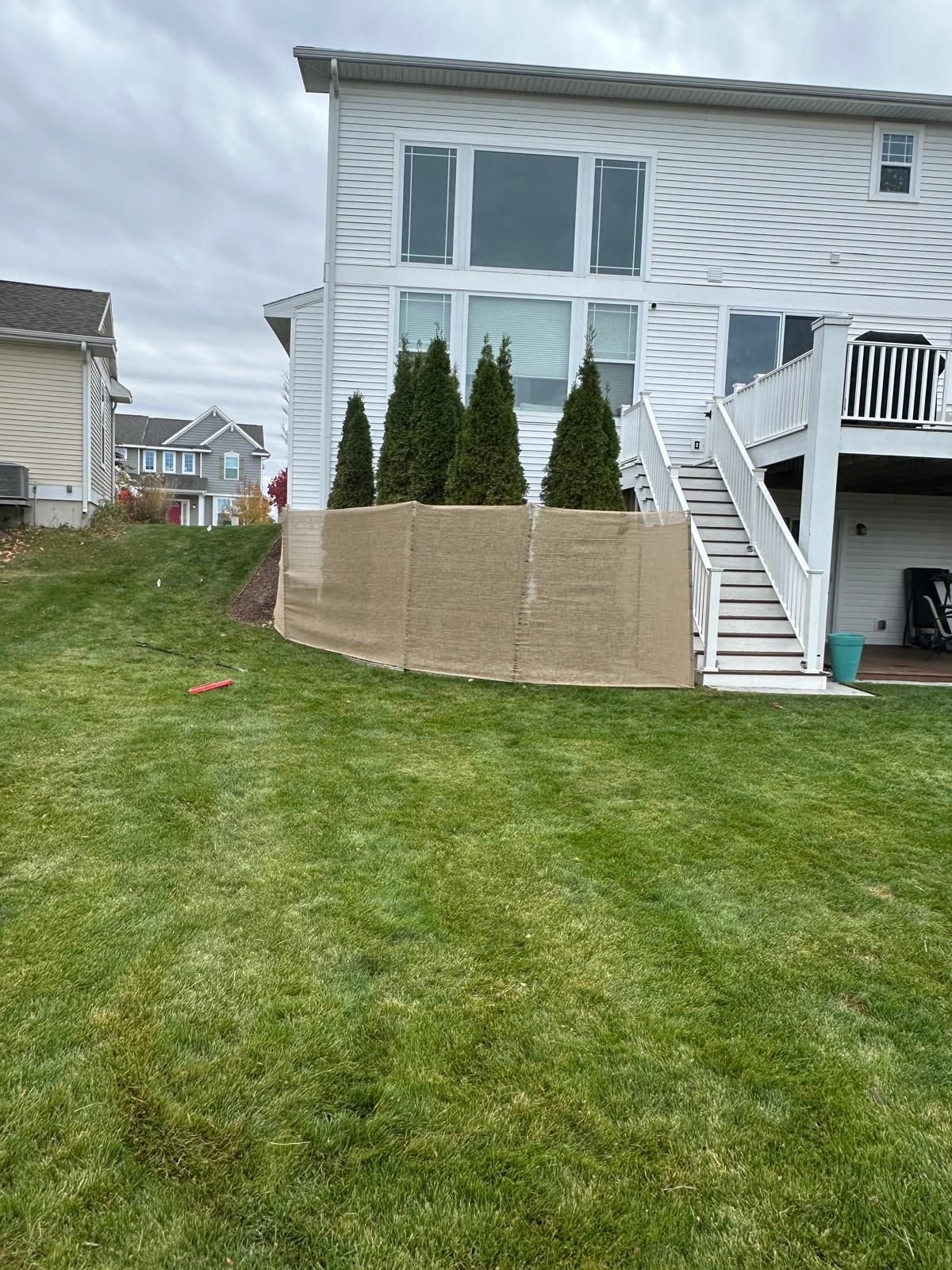 A white house with a deck and stairs features a row of evergreen trees wrapped in burlap along the base.