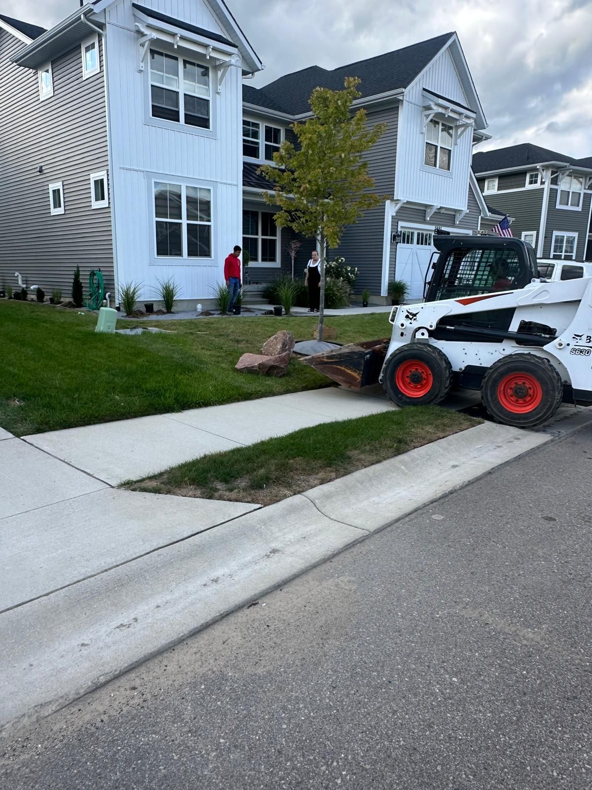 A skid-steer loader sits in a suburban front yard next to a young tree and two people near a white house.