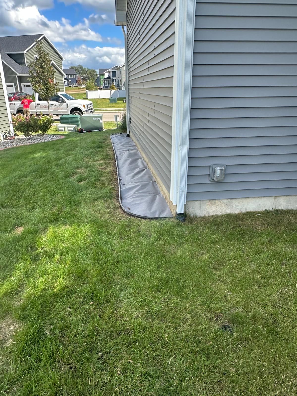 A gray plastic landscape barrier runs along the foundation of a gray-sided house, situated next to a lawn and driveway.