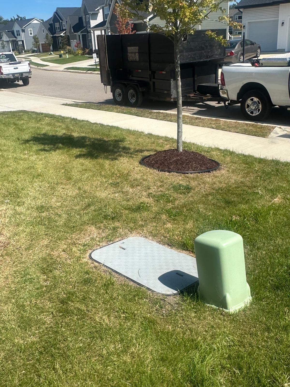 A utility box and access cover on a residential lawn, with a trailer and truck parked on the street in the background.