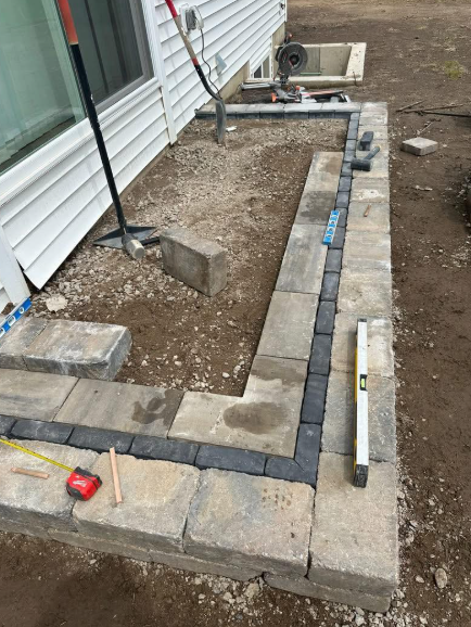 Construction of a retaining wall against a white house. Grey pavers are being laid on gravel.