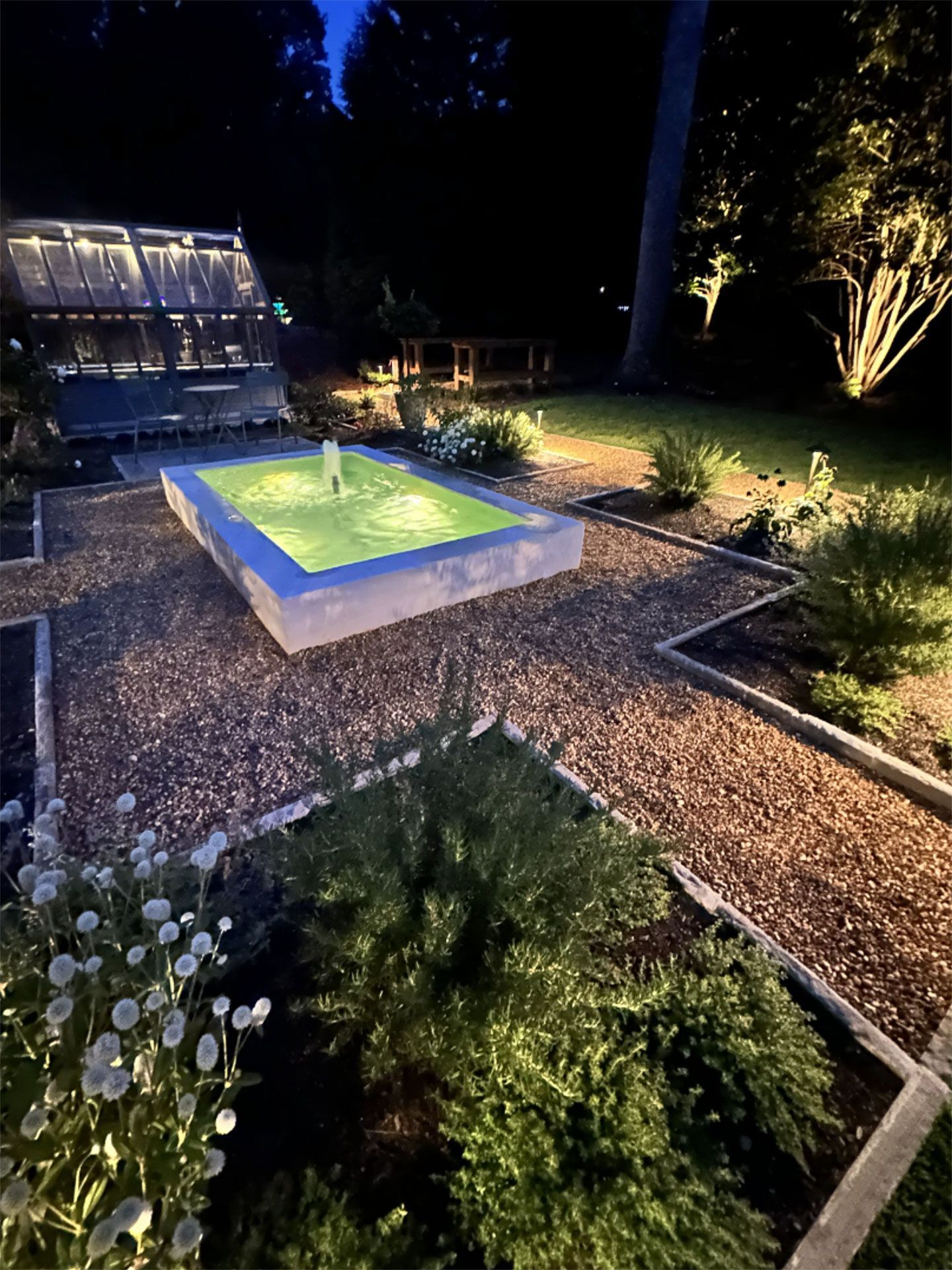 A night garden with a lit fountain, pathways, and illuminated plants under a dark sky.