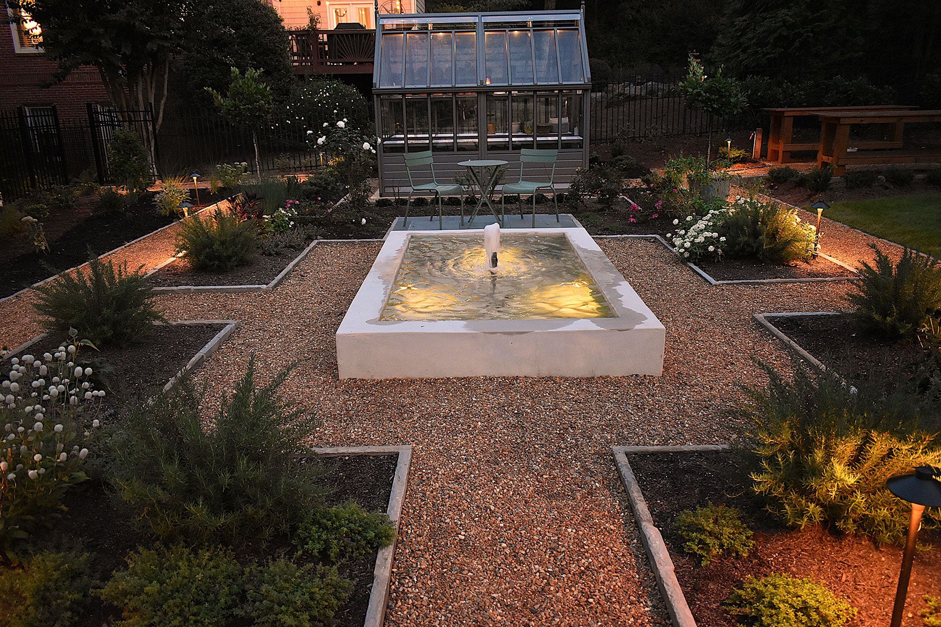 A garden with a fountain, gravel paths, and a greenhouse, illuminated by warm lighting.