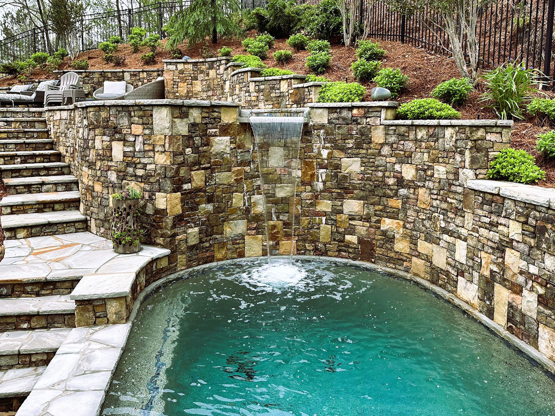 Stone-walled pool with waterfall feature, steps, and terraced landscaping against a hillside.