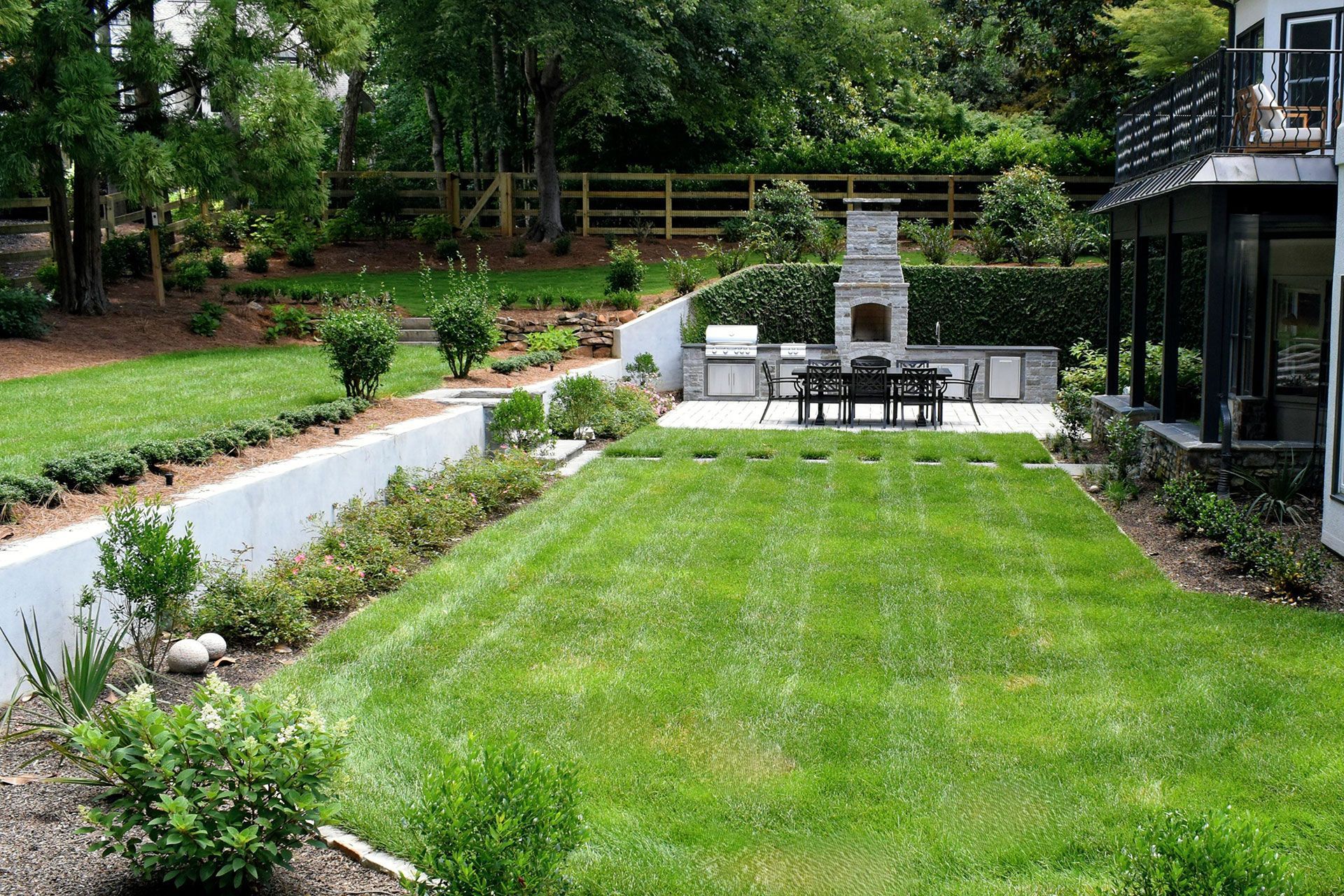 Well-manicured backyard with lawn, stone patio, outdoor kitchen, fireplace, and lush greenery.