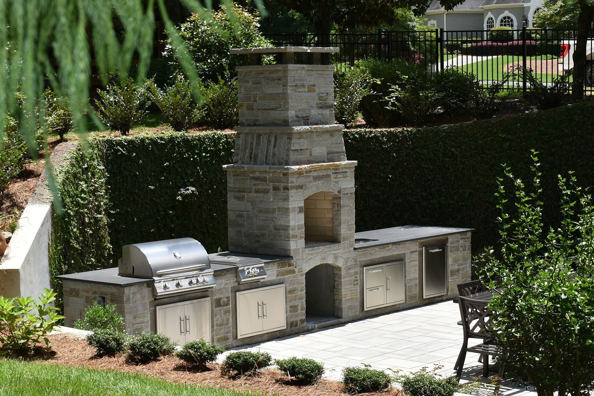 Outdoor stone kitchen with grill, oven, and storage, set against a green hedge.